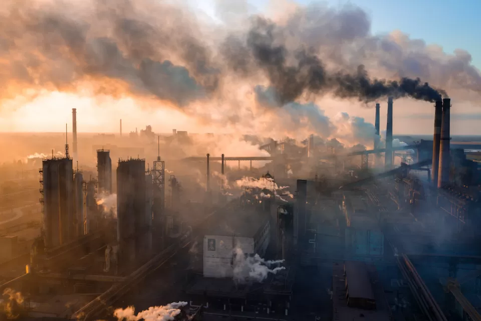 Aerial view of industrial plant with large smokestacks, surrounded by smoke with sunset sky in background.